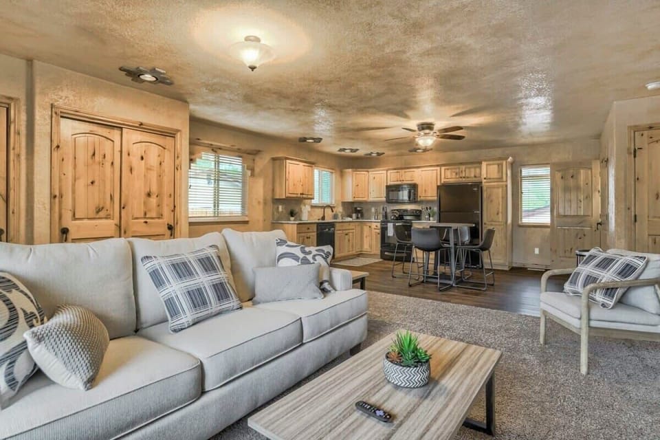 Open living area where the sofa and coffee table flow naturally into the kitchen beyond. Ceiling fans, warm wall tones, and wood cabinetry create a connected space where guests can lounge, chat, and gather.