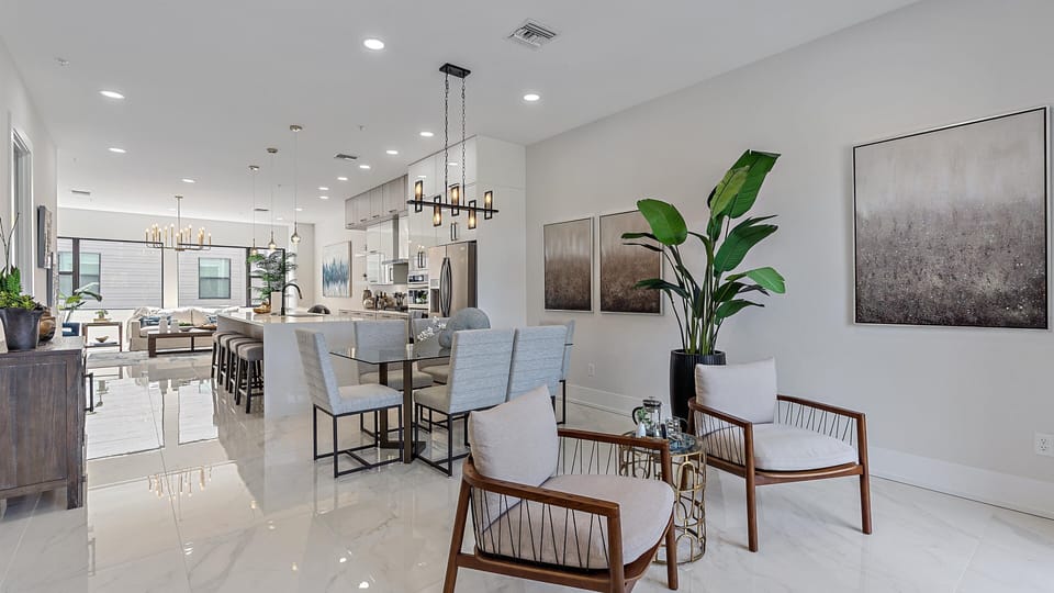 A modern open-concept living and dining area with light gray walls, marble floors, and contemporary furniture, including a dining table, lounge chairs, and a large plant.
