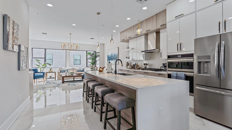 Modern kitchen with a large island and wooden barstools. There are stainless steel appliances, white cabinets, and pendant lights. The kitchen opens into a spacious living area with large windows.
