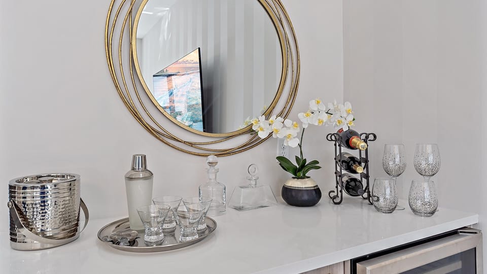 A drink station featuring a round mirror, flowers, a wine rack with bottles, cocktail glasses, decanters, a shaker, and an ice bucket on a white counter.