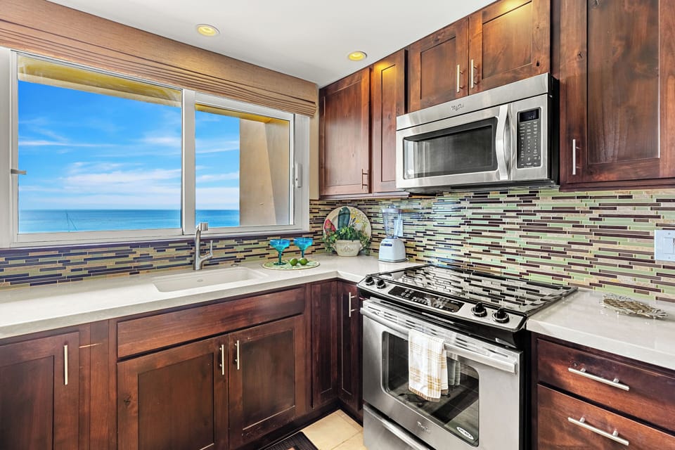 Prepare your favorite meals in the fully equipped kitchen featuring modern appliances, rich wood cabinetry, and scenic ocean views right from the window.