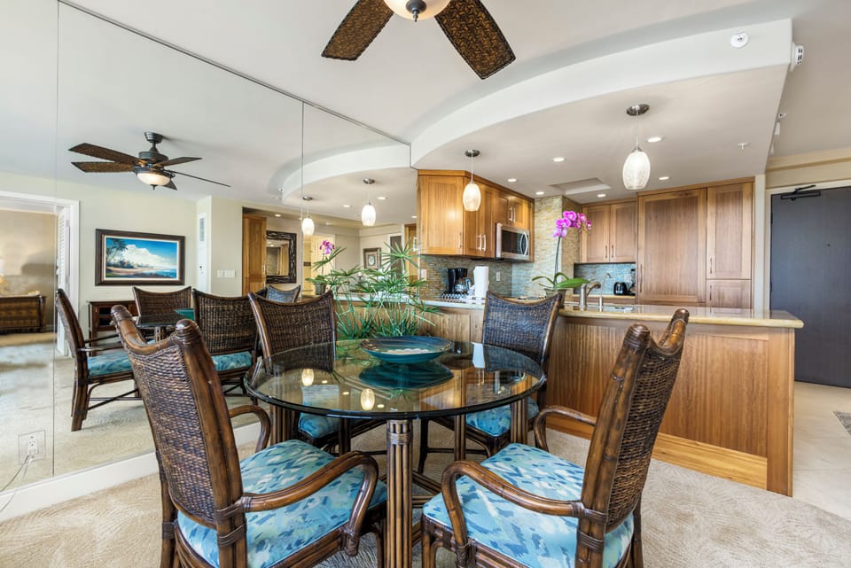 Gather around the elegant dining table where memorable meals await, flowing seamlessly into your fully-equipped kitchen with modern appliances, warm wood cabinetry, and tropical island charm.