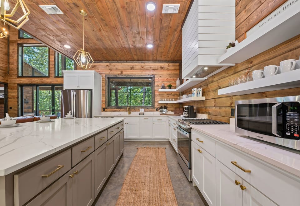 Awesome view inside the kitchen showing white cabinetry, marble countertops, and wooden shelves against a backdrop of wooden walls and a large window.