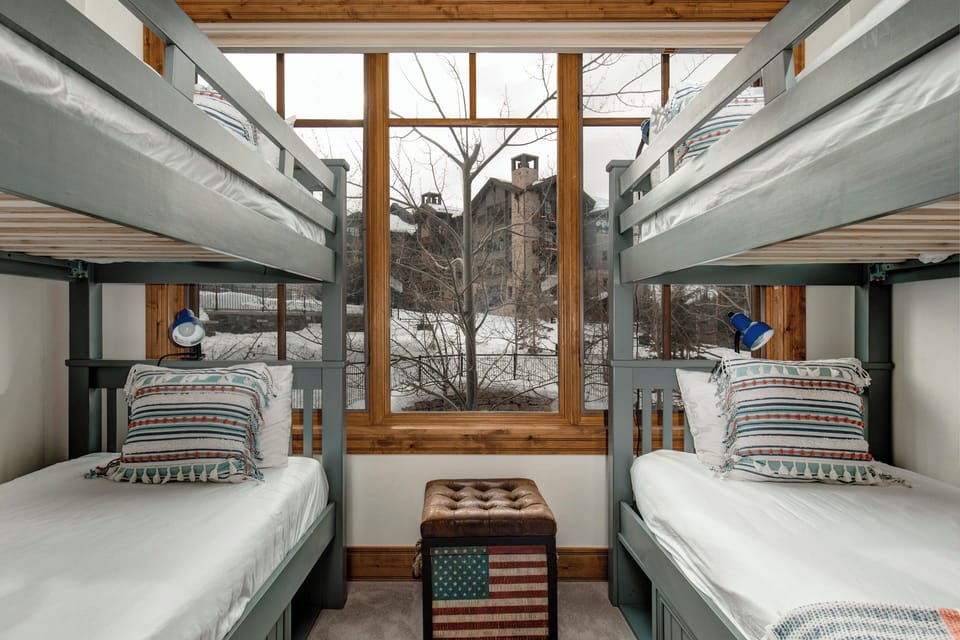 - Two twin over twin bunk beds offer a fun & functional sleep setup for kids or groups
- Oversized window frames a scenic view of snowy rooftops & bare winter trees
- Vintage style ottoman with a patriotic print adds charm & extra seating/storage