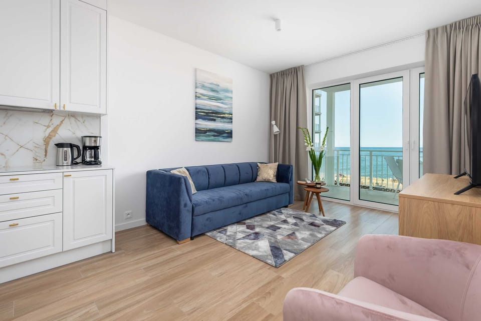 A bright living room with a blue sofa, a glass coffee table, and a balcony with a sea view.