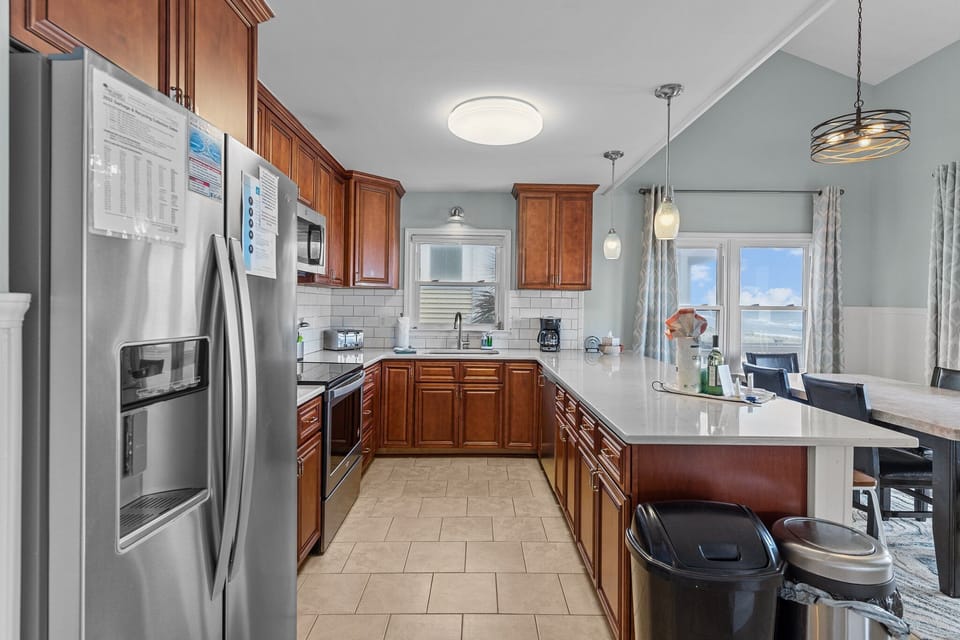 Kitchen with warm wood cabinets, white quartz bar seating, stainless appliances, subway tile backsplash, pendant lights, and coffee station.
