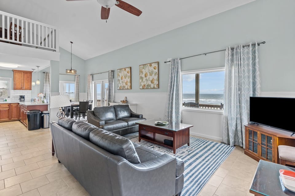Bright living room with vaulted ceilings, leather seating, coastal art, and windows that bring in natural light and beautiful ocean views.