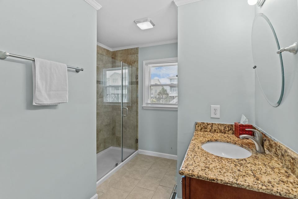 Bathroom with a granite vanity, round mirror, bright window, tiled floors, and a glass walk-in shower with neutral coastal finishes.
