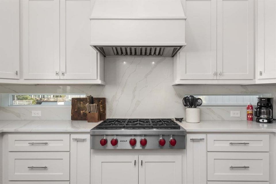 Chef-style kitchen range with stainless hood, white cabinetry, marble-look backsplash, and modern gas cooktop.