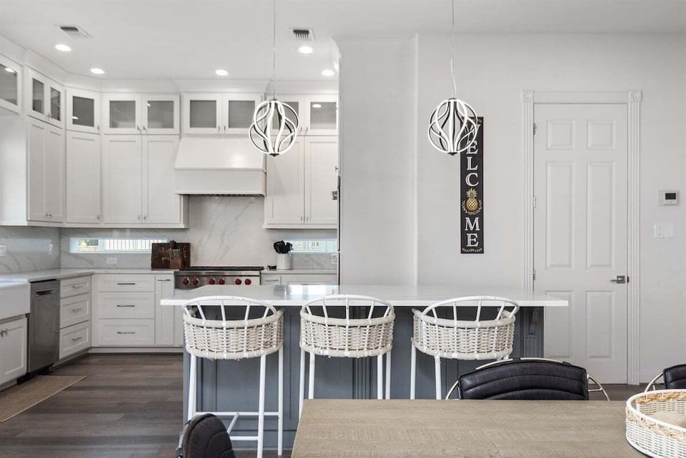 Kitchen island with bar stools, pendant lighting, white cabinets, stainless appliances, and coastal welcome decor.