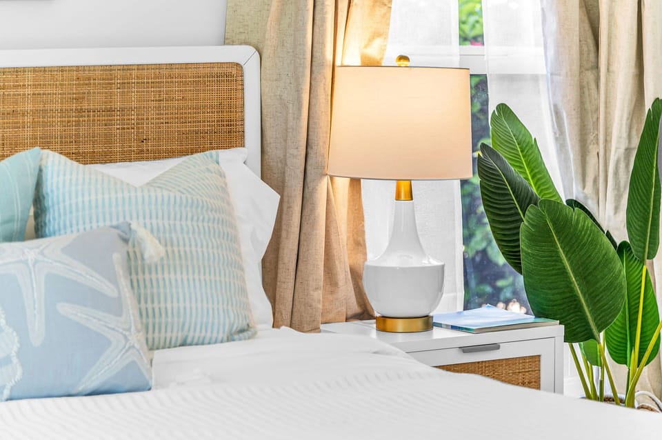 The primary bedroom suite is coastal "Serena & Lily" inspired. The king bed is a mango wood platform bed with a woven cane head and footboard in a natural finish, topped with a LUCID aloe gel memory foam mattress and 100% luxurious cotton linens.