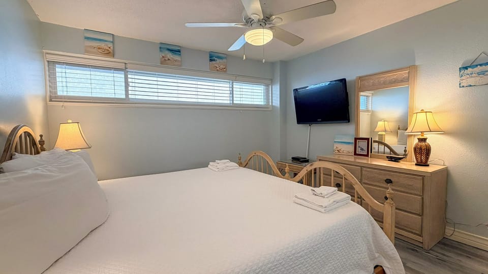 Relax in your cozy bedroom with coastal charm, featuring fresh linens, a ceiling fan for comfort, and cheerful beach-themed decor.