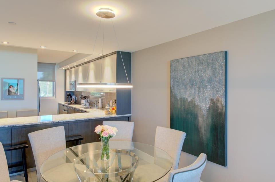 Glass-top dining table surrounded by beige upholstered chairs and a large abstract blue-green painting.