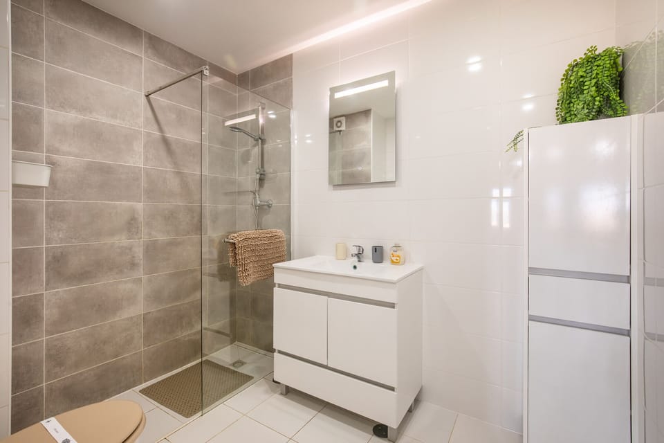 A modern and bright bathroom featuring a shower area separated by a glass screen and lined with large grey tiles. The rest of the space has white glossy tiles and includes a white vanity unit with under-storage and a tall white cabinet topped with a decorative plant.
