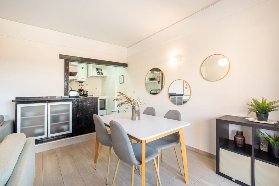 A bright and modern dining area that flows into an open kitchen space. The dining section features a clean white table with wooden legs and four stylish grey chairs. The wall is decorated with an attractive cluster of three round, gold-framed mirrors. A convenient bar area with black cabinetry and frosted glass doors visually separates the space from the kitchen.