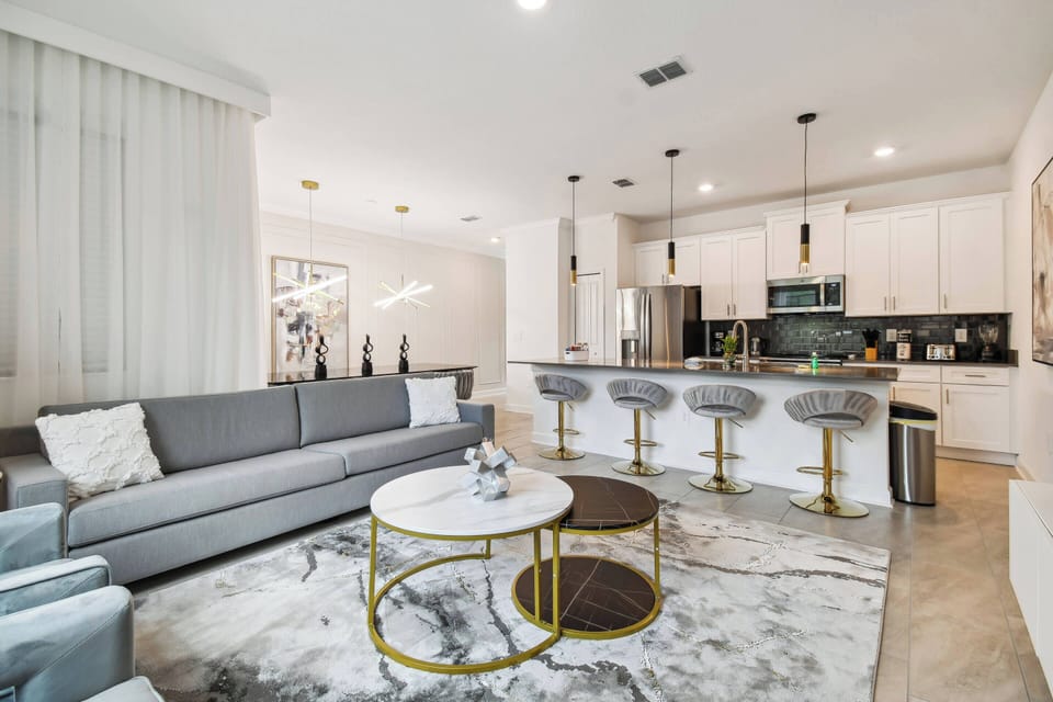 Relax in a stylish open-concept living area with a plush gray sofa and elegant gold-accented coffee tables. The modern kitchen, featuring stainless steel appliances and bar seating, is ideal for cooking and socializing.