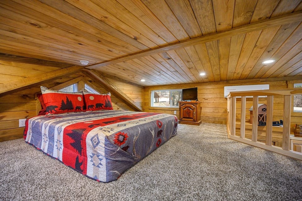 A cozy loft bedroom with a wooden ceiling and walls, carpeted floor, a bed with a bear-themed quilt, a small window, a TV, and a wooden railing by the stairs.