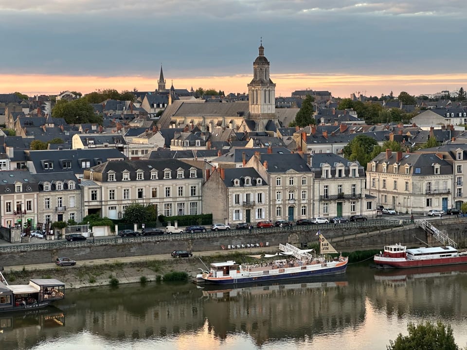 The Angers skyline at sunset, with moored Bar à Vin barges along the quayside and the city's historic rooftops and church towers reflected in the Maine River.