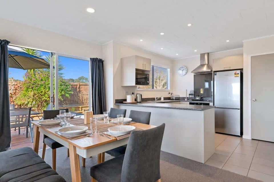 Modern open-plan dining area with timber table, six chairs and stainless kitchen — family-friendly Rotorua holiday home