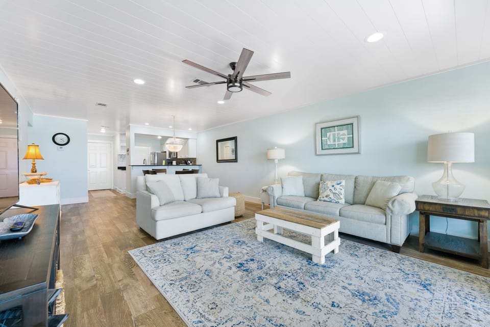 Sink into plush seating in your bright, coastal-inspired living space where gentle ceiling fans and soft lighting create the perfect atmosphere for relaxation.