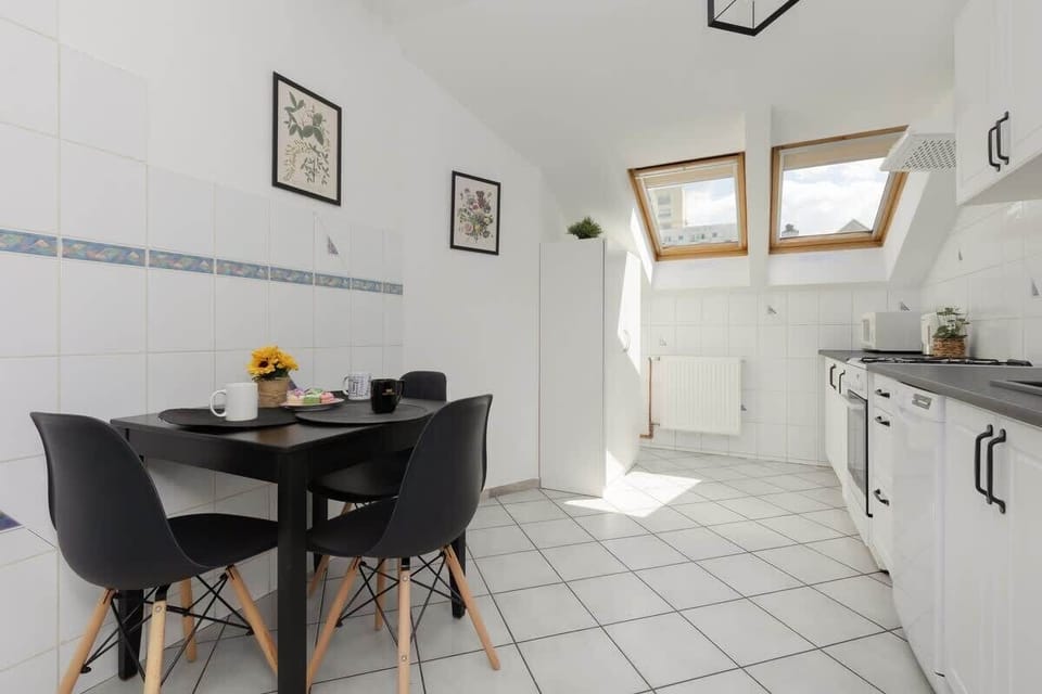 A fully tiled white kitchen equipped with modern appliances, black dining chairs, and abundant natural light from skylights.

