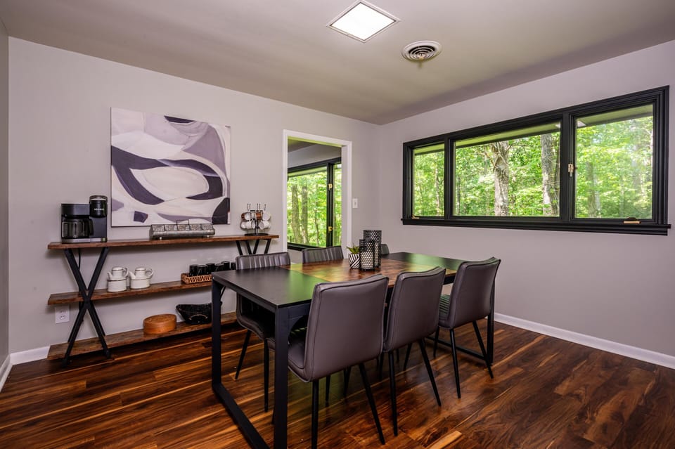 The dining room features a wood-top table with six leather chairs, overlooking forest through wide black-trimmed windows.