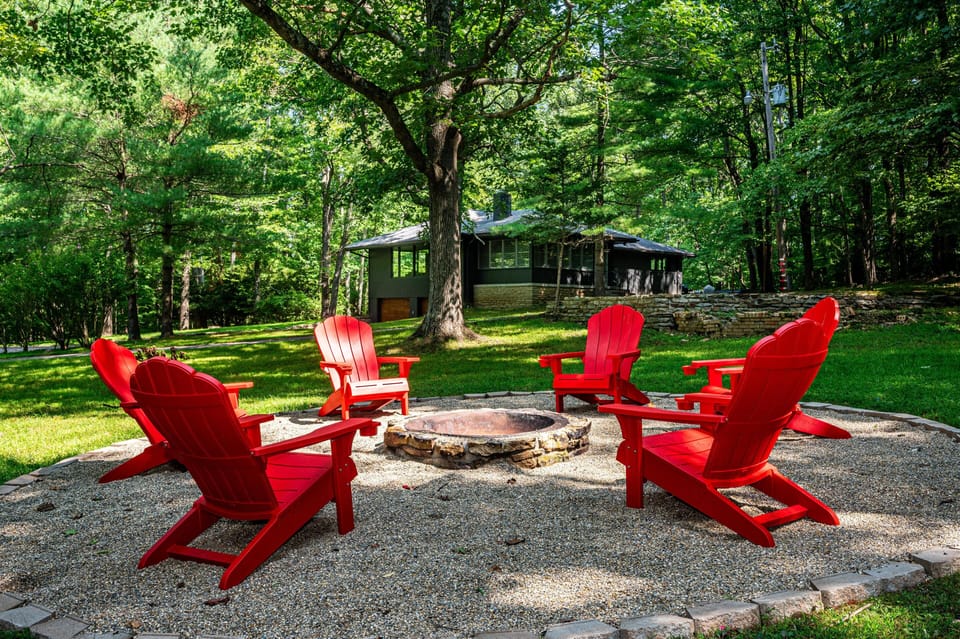 Six red Adirondack chairs encircle a stone fire pit on gravel, with a green house and trees in the background.