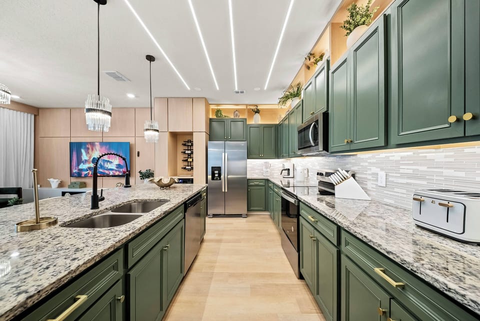 Experience a chic kitchen featuring deep green cabinets and elegant gold hardware that beautifully contrasts with light granite countertops. Well-lit by modern fixtures, this space is perfect for meal prep or casual breakfasts.