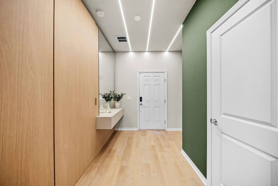 Step into a bright entryway featuring sleek wooden cabinetry and a soothing green accent wall that welcomes you. The warm wooden flooring and modern lighting create a clean, airy atmosphere, inviting you to explore further.