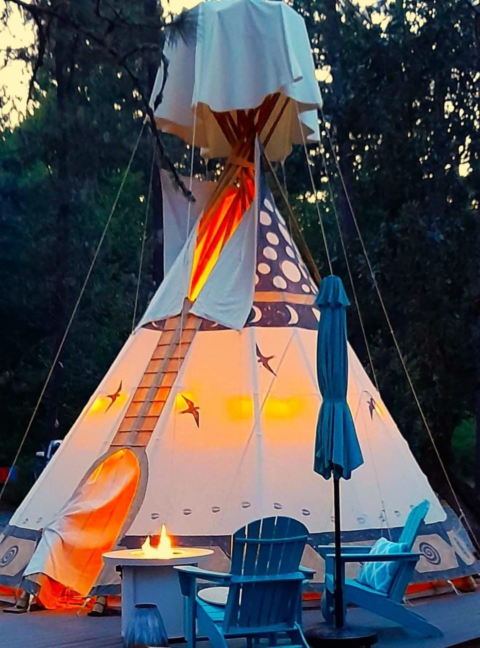 The Sky Spirit is an 18-foot Tipi on an oversized deck. It has outdoor chairs, a large umbrella, a propane fire pit: a picnic table, and a briquet BBQ grill.