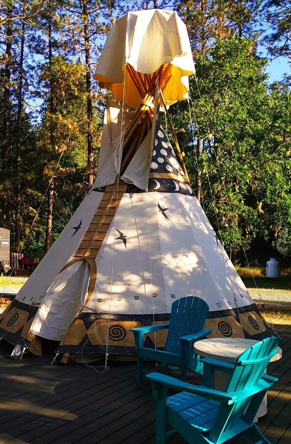 The Sky Spirit is an 18-foot Tipi on an oversized deck. It has outdoor chairs, a large umbrella, a propane fire pit: a picnic table, and a briquet BBQ grill.