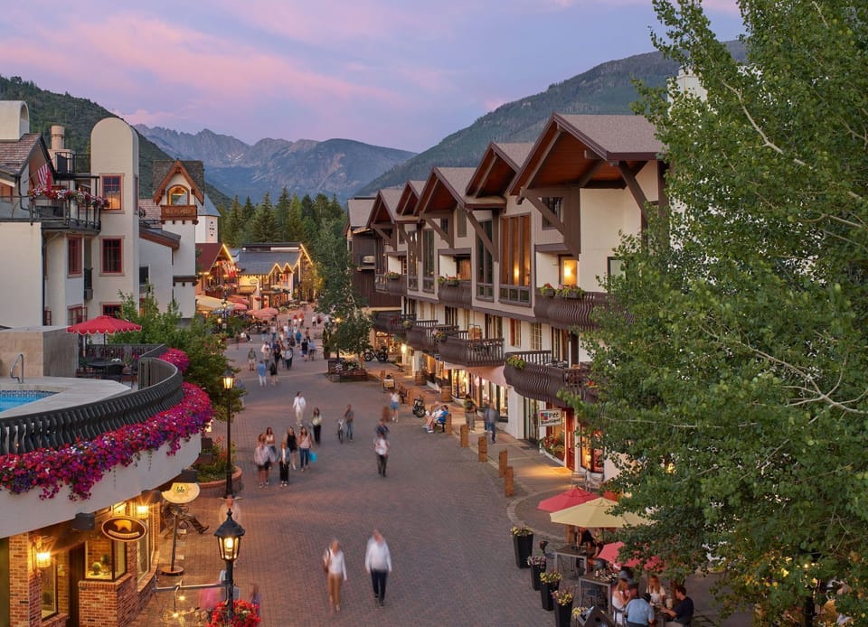 Brick promenade lined with flowering balconies fills with pedestrians beneath a pink evening sky