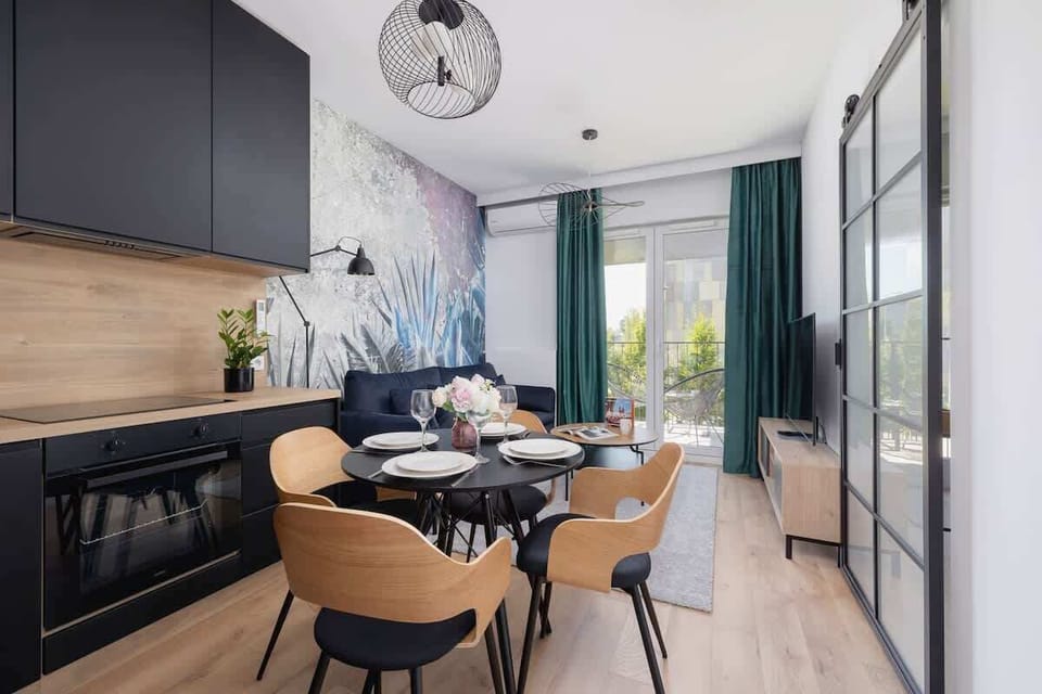 An open-concept kitchen and dining area with black cabinetry above and wood-finish cabinets below. A round dining table with contemporary chairs sits beneath a chic pendant light.
