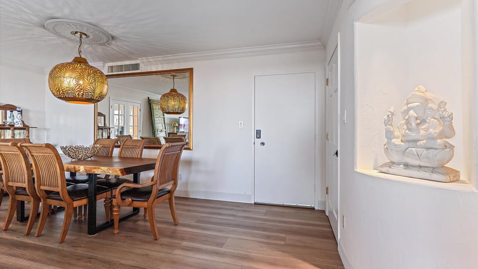A dining room with a wooden table and chairs, pendant lights with decorative shades, a large wall mirror, and a niche displaying a white statue. The walls and floor are different shades of wood.
