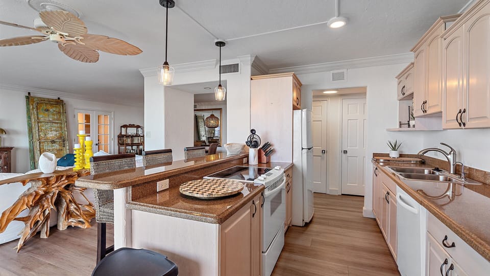 A bright, open-plan kitchen with wooden countertops, white cabinets, a ceiling fan, and pendant lights. There is a bar counter with seating and a view of the adjacent dining area.