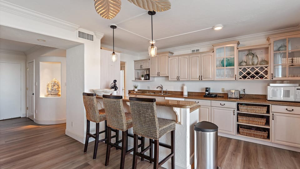 A modern kitchen with light wood cabinetry, a granite countertop, three wicker bar stools, pendant lighting, and stainless steel appliances. An entryway with a sculpture is visible on the left.