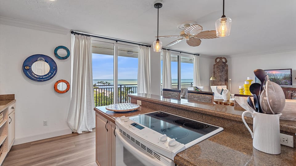 Modern kitchen with a view of the ocean, featuring a stove, countertop, utensils in a holder, and various wall decorations. Sliding doors open to a balcony with a scenic seascape.