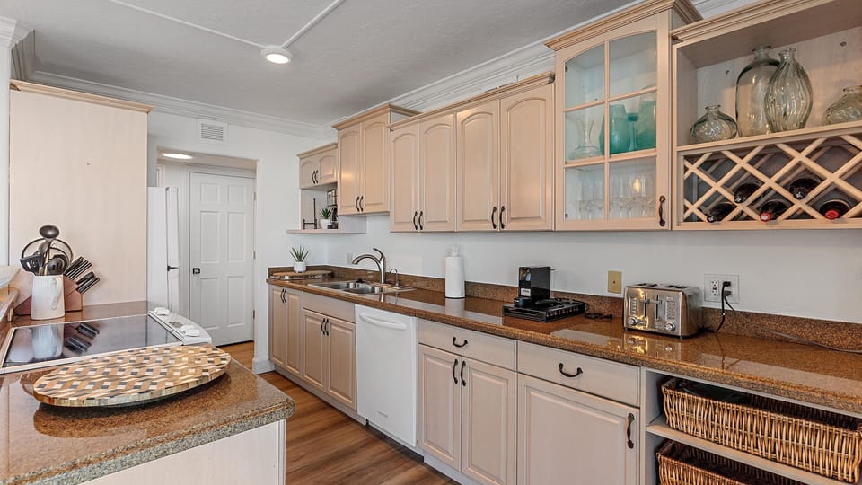 A modern kitchen with beige cabinetry, granite countertops, a white dishwasher, electric stove, various kitchen items, and glass cabinet doors. A wine rack is built into the cabinetry.