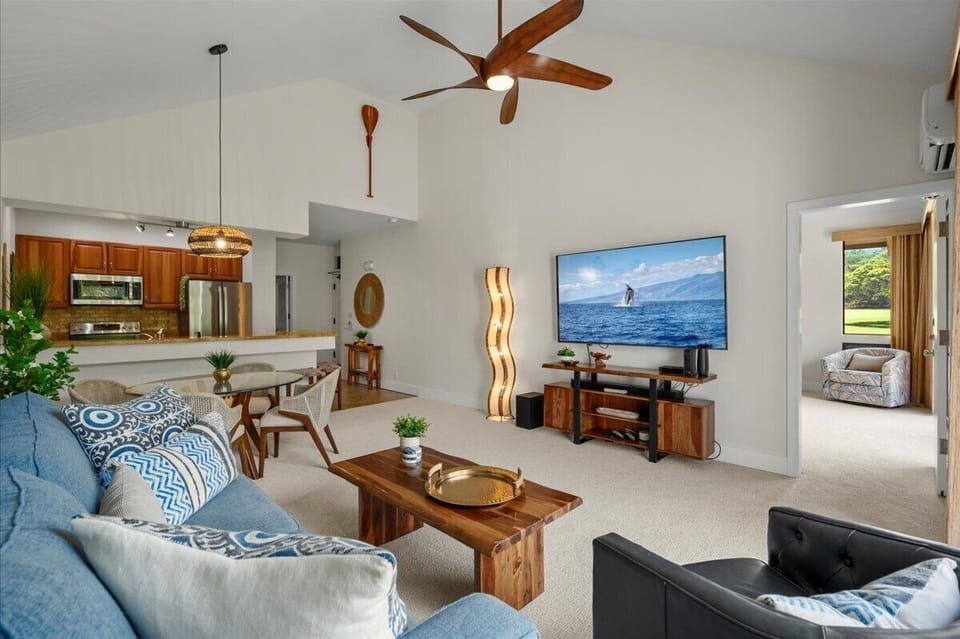 Relax in your bright, open-plan living area with a large TV, stylish furniture, and a fully-equipped kitchen. High ceilings and tropical decor invite you to unwind in comfort.