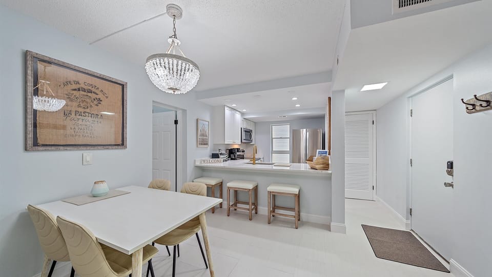 A dining area and kitchen with an open layout. The room features a white dining table and a breakfast bar with stools.