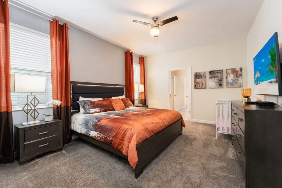 King bedroom with rust-orange bedding, tall sunlit windows, a beach-themed TV, and a crib for family-friendly stays.