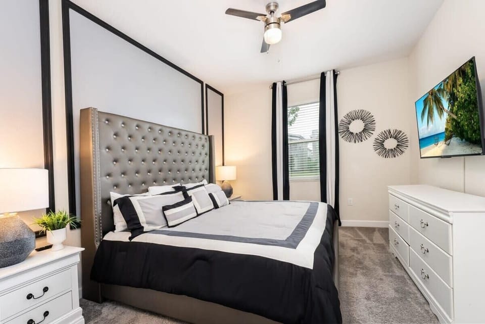 Private king bedroom with a tufted gray headboard, crisp black-and-white bedding, and a wall-mounted TV displaying a tropical beach scene.