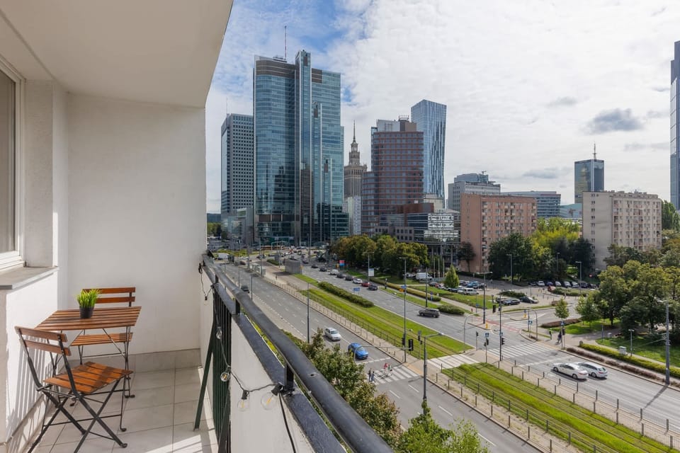 A balcony overlooking a bustling city street with tall skyscrapers and green landscaping. The space features a small table and chairs, ideal for relaxing while enjoying the view.
