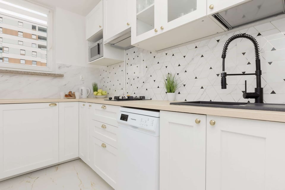 A modern white kitchen with a black faucet and a striking monochrome backsplash. White cabinets and countertops create a crisp, open feel.
