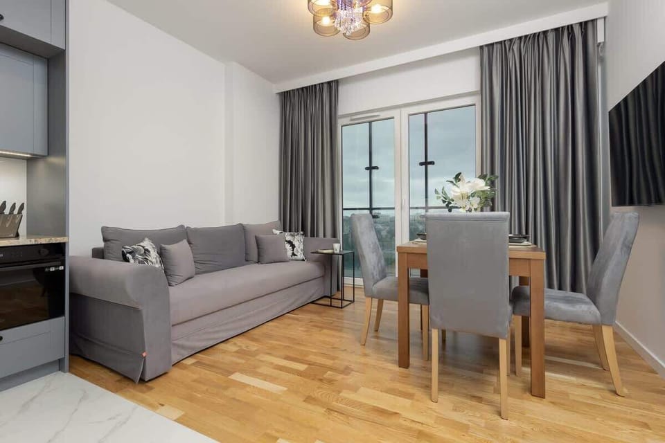 An open-plan living and dining area with a gray sofa, wooden floors, and a dining table with seating for four near large windows.

