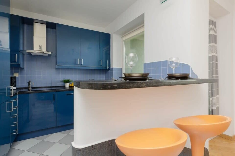 A closer look at the kitchen and bar area with deep blue cabinets, an L-shaped countertop, and bright orange bar stools adding a fun accent.

