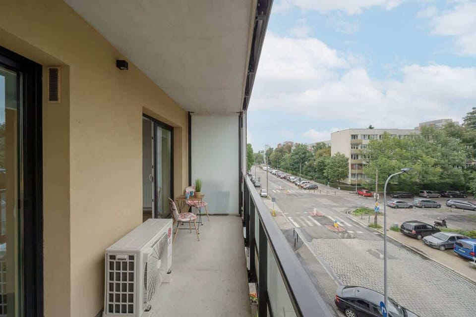 A balcony with a view of a quiet street, featuring a small table, potted plants, and a railing for a relaxing outdoor space.

