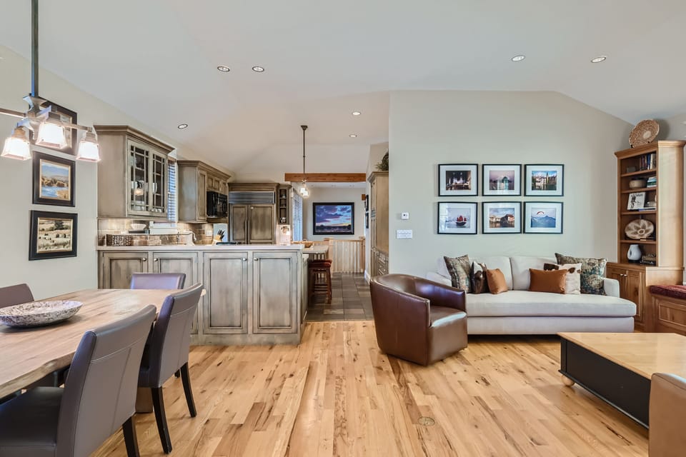 A modern open-concept living area featuring a kitchen with wooden cabinets, a dining table with chairs, a couch, and a wall adorned with framed photos. The room has hardwood floors and recessed lighting.
