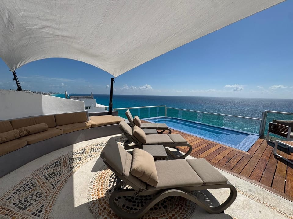 Curved stone bench with tan cushions under a white shade sail, facing four cushioned lounge chairs by a glass-railed infinity pool.