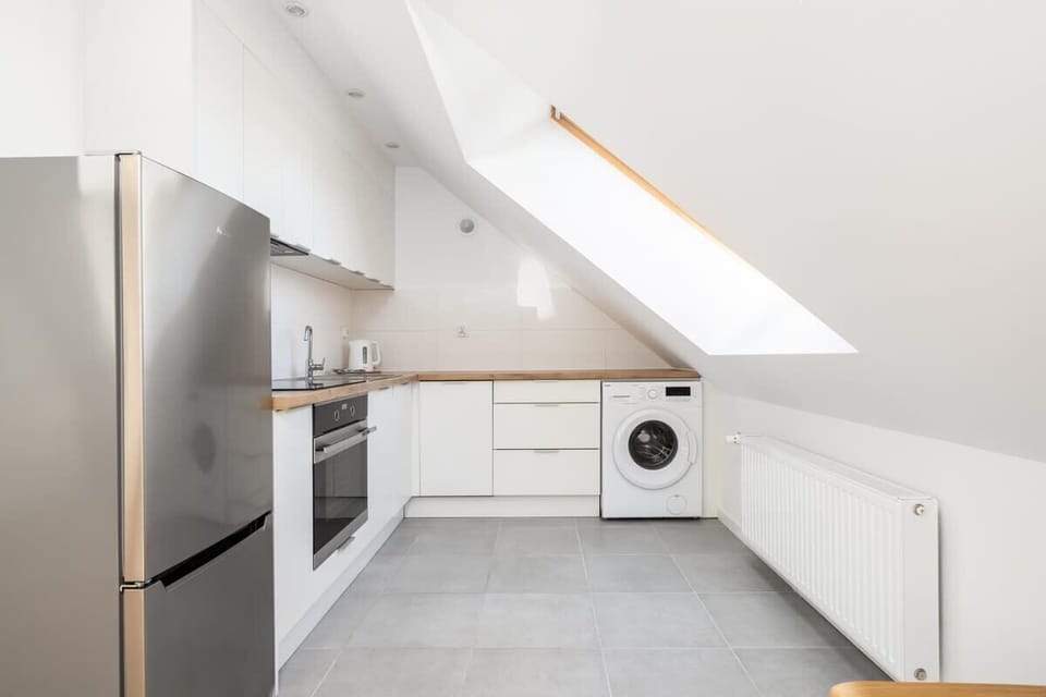 A modern kitchen in a compact attic space, featuring white cabinets, stainless steel appliances, and slanted ceilings.

